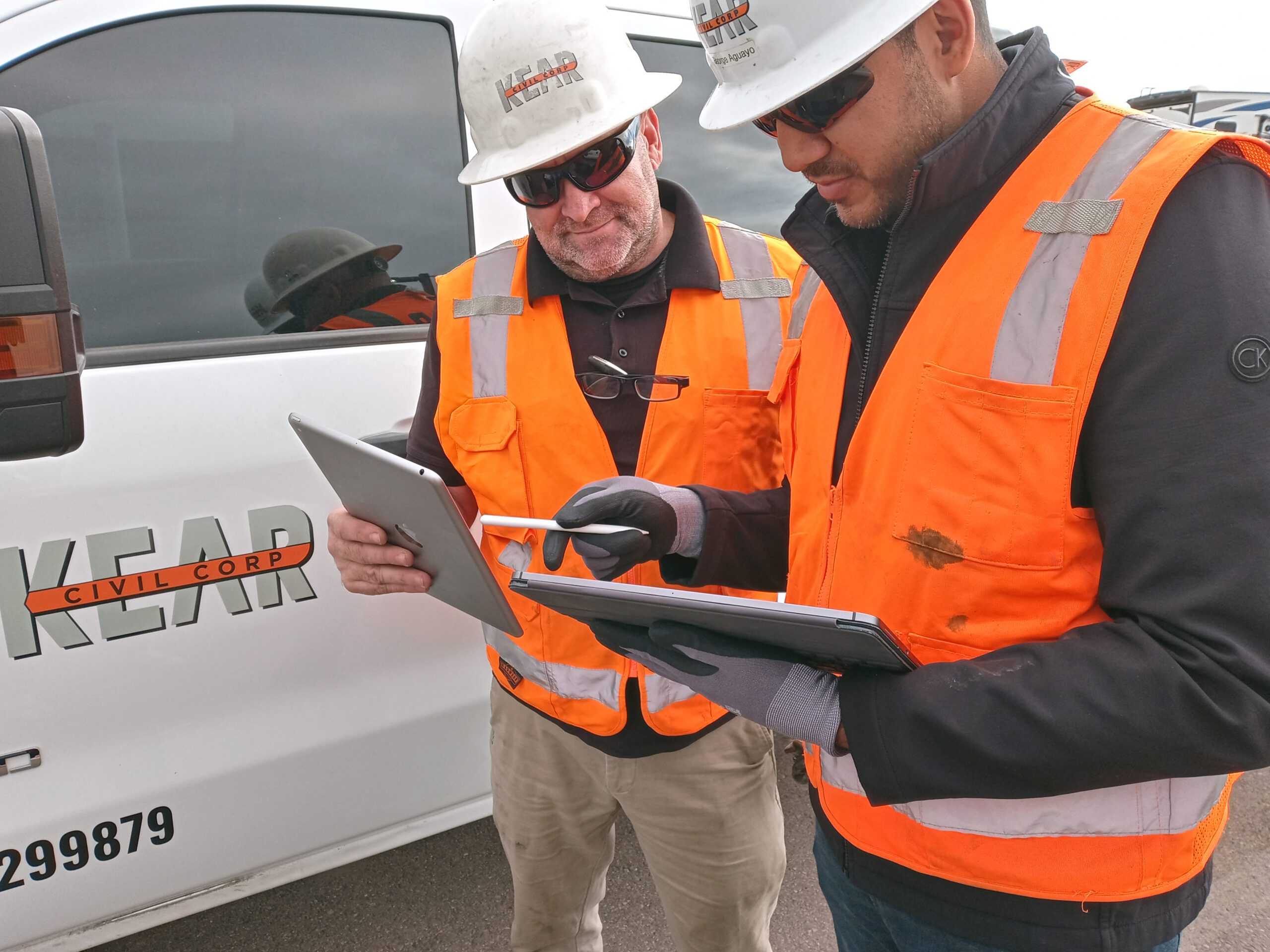 Two construction workers in orange vests and helmets looking at a tablet near a home improvement company van.