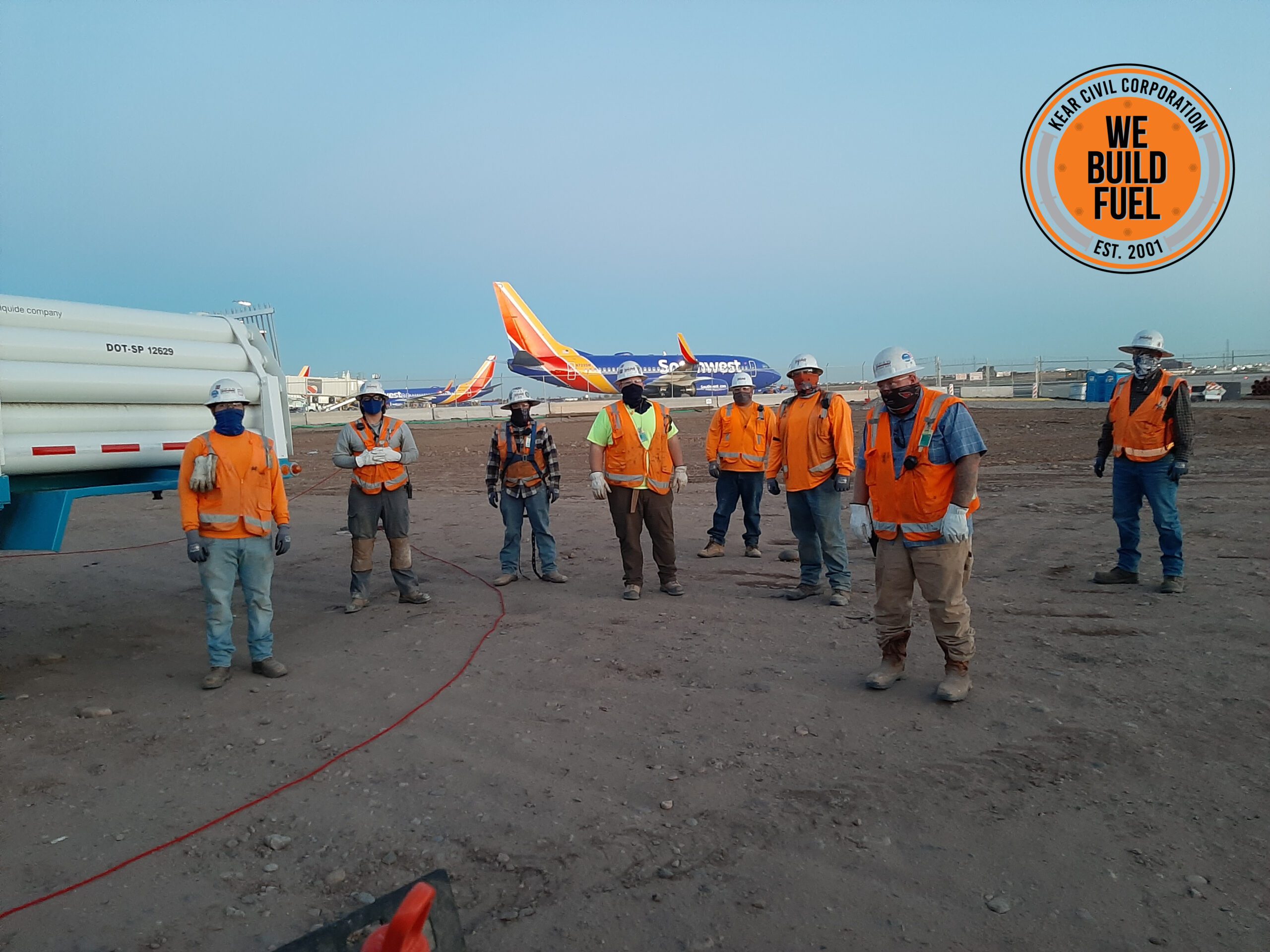 A group of construction workers in high-visibility vests and hard hats standing at a worksite with a truck and airplane in the background, building a new home.