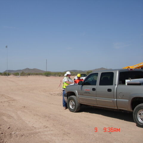 Two construction workers in reflective vests discussing about a project beside a pickup truck on a vast, barren construction site with clear skies.
