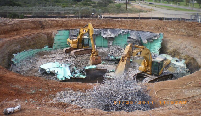 About excavators working in a large pit with debris and twisted metal, surrounded by a dirt embankment.
