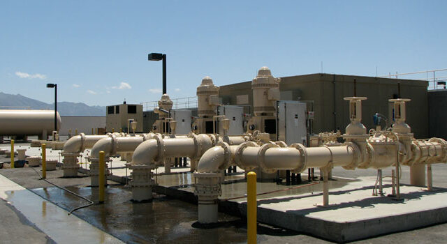 An industrial site featuring pipelines and a pump station, with various machinery on concrete bases, under a clear sky and distant mountains in the background.