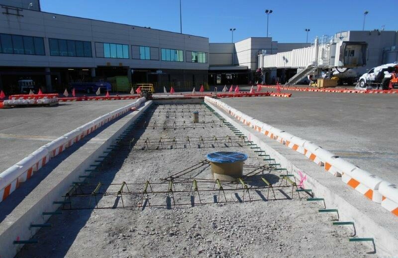 Construction area with rebar framework set up in a trench for the Fuel Hydrant Relocation, Nashville International Airport terminal and new PBBs in the background under a clear sky.