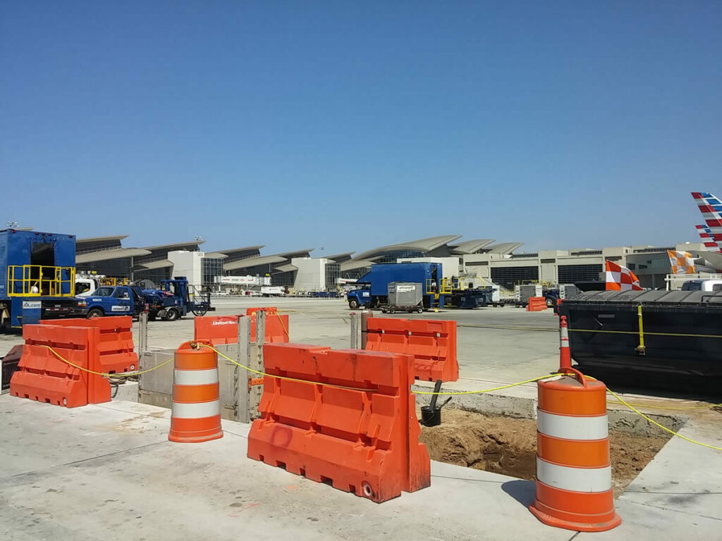 Construction barriers and cones at an airport tarmac with ground service vehicles and airplanes visible in the background near Hydrant Fuel Pits under a clear blue sky.