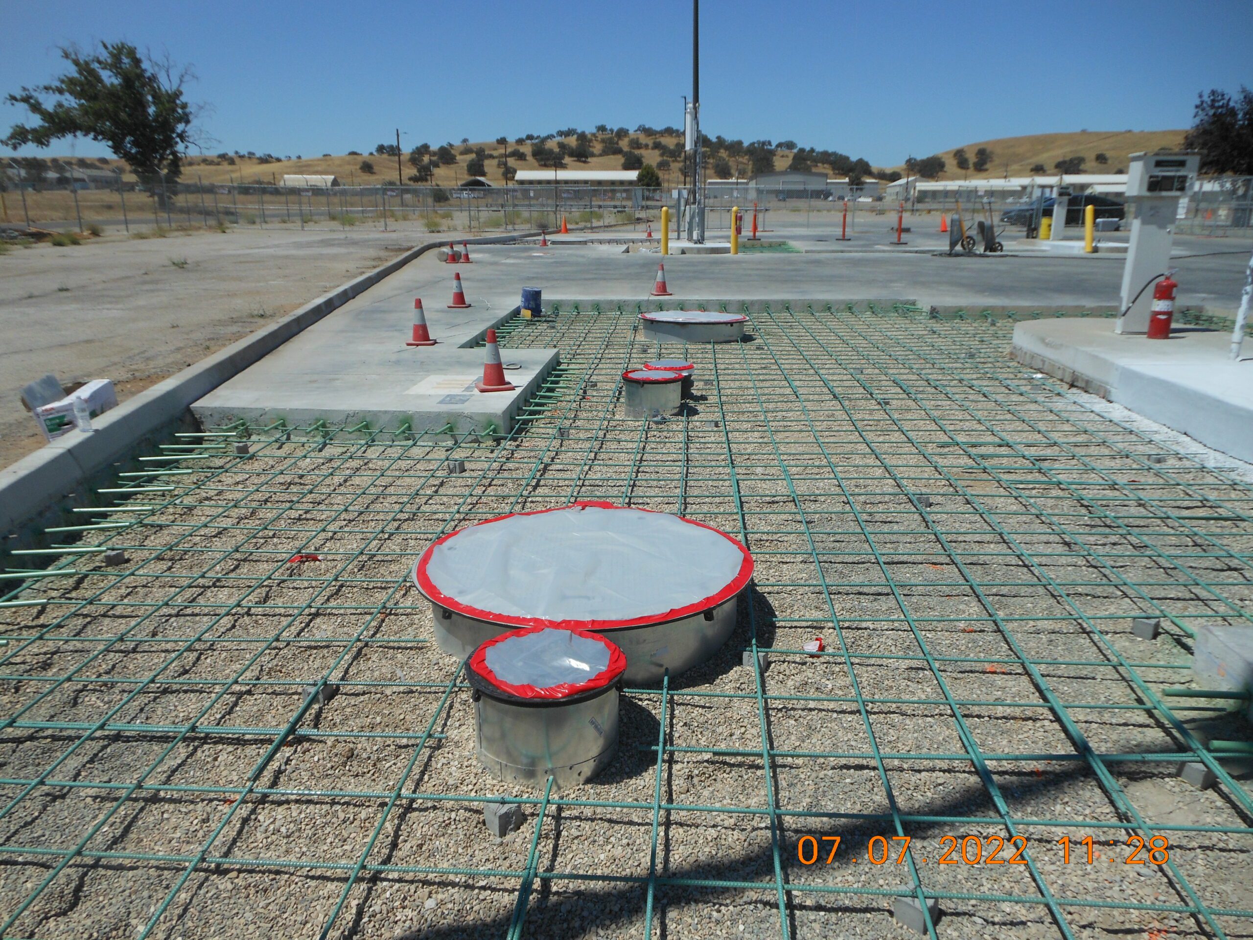 Construction site featuring an unfinished concrete base with exposed steel reinforcements and circular structures, set against a hilly background under a clear sky.