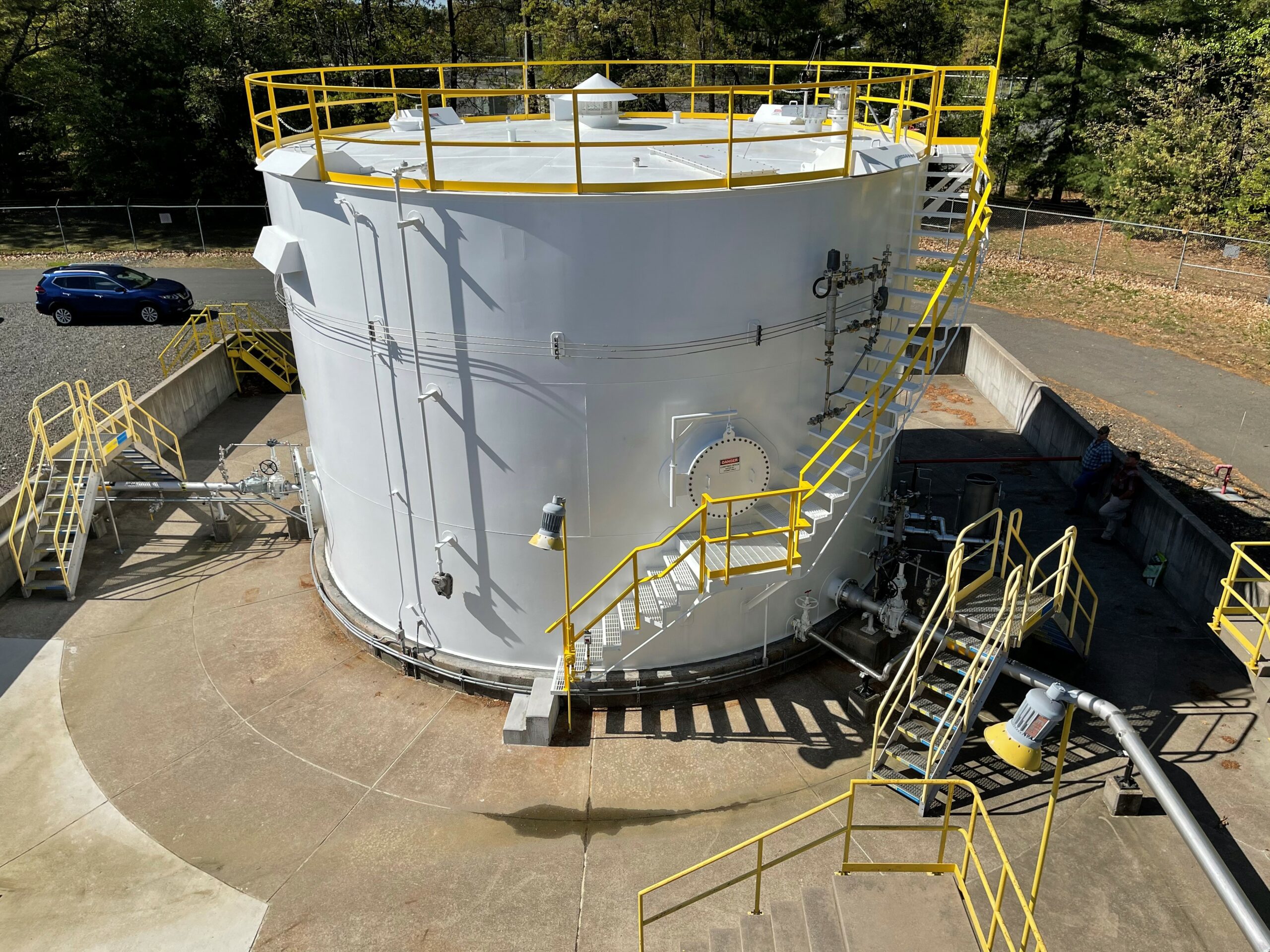 Large industrial storage tank with white exterior and yellow railings, managed by the US Army Corps of Engineers, surrounded by safety stairs and concrete platform, with a car parked nearby.