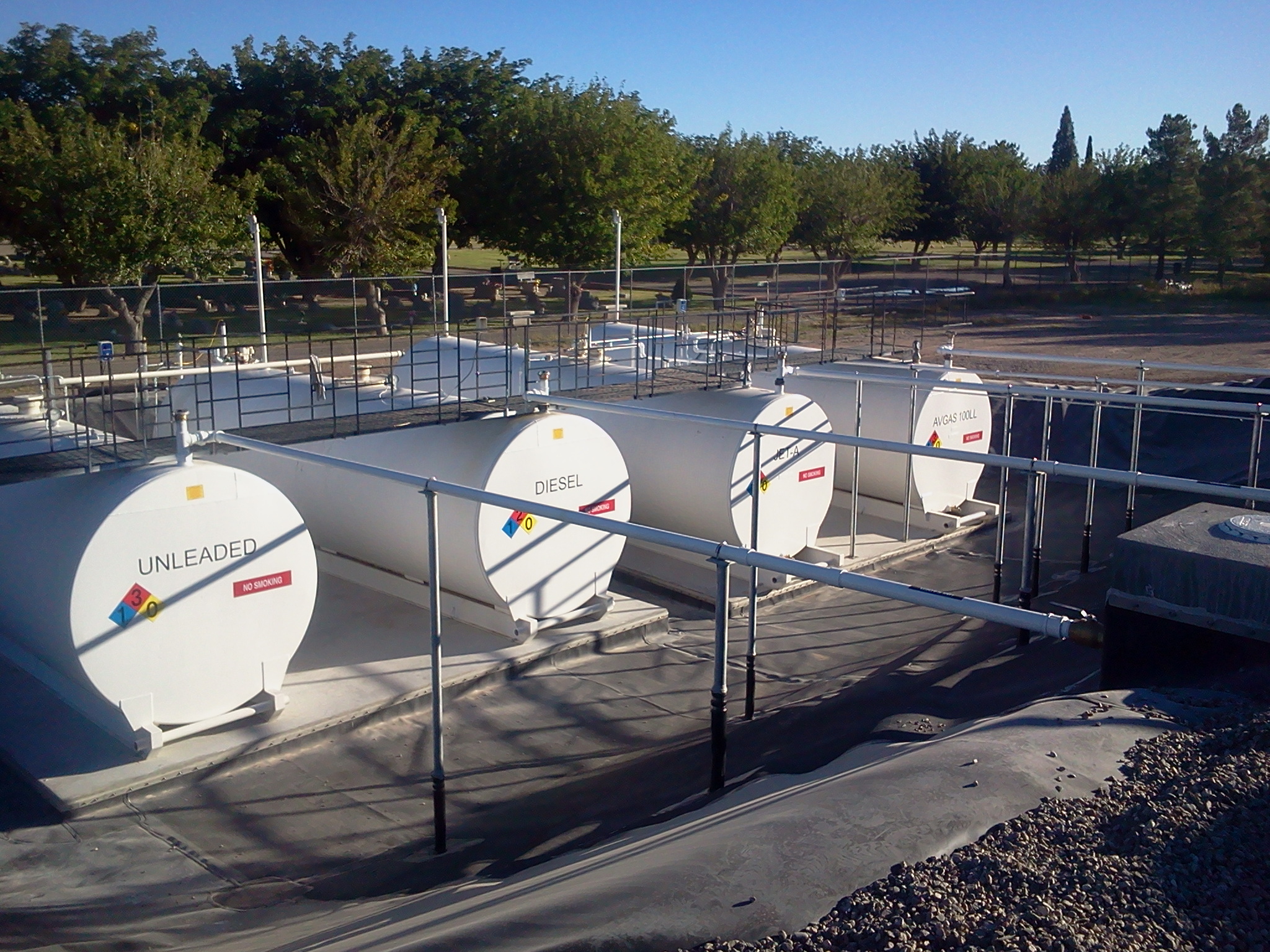 A fuel farm in Deming, New Mexico, with three labeled tanks for unleaded and diesel fuels, surrounded by a metal fence, in a sunny outdoor setting.