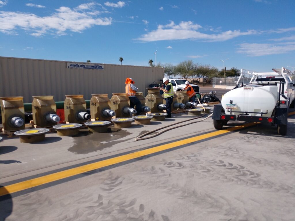 Workers in high-vis vests inspecting hydrant fuel pits parts laid out on the ground next to a white tanker truck under a clear sky at Phoenix Terminal 3.