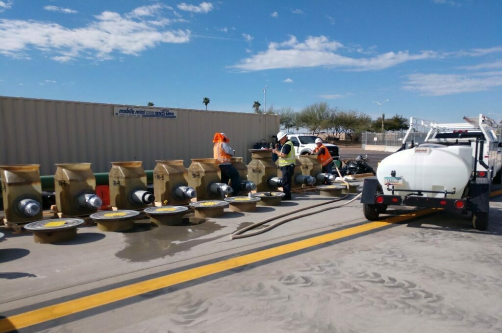 Workers in high-vis vests inspecting hydrant fuel pits parts laid out on the ground next to a white tanker truck under a clear sky at Phoenix Terminal 3.