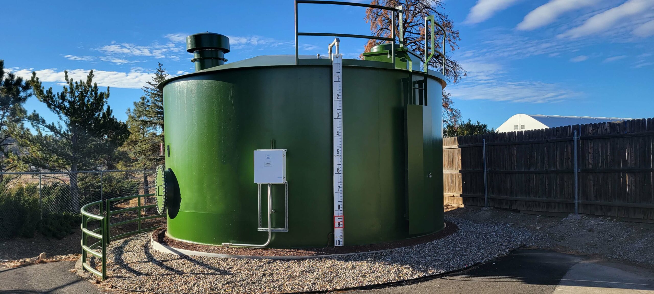 Large green industrial water tank with a level gauge and a smaller attached cylinder, situated in a fenced area with a clear blue sky, maintained by the City of Flagstaff.