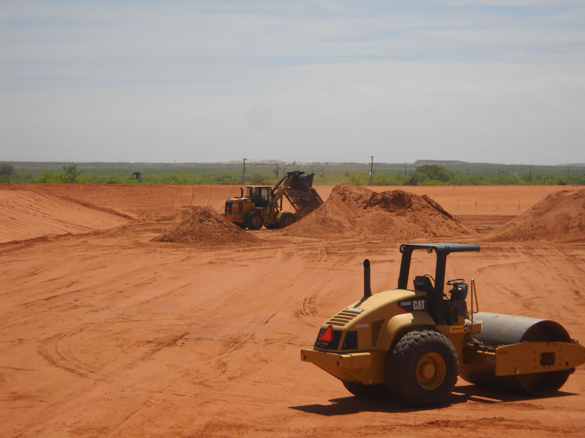 Two construction vehicles, a bulldozer and a compactor operated by the U.S. Army Corps of Engineers, working on dirt mounds in a large open field under a clear sky.