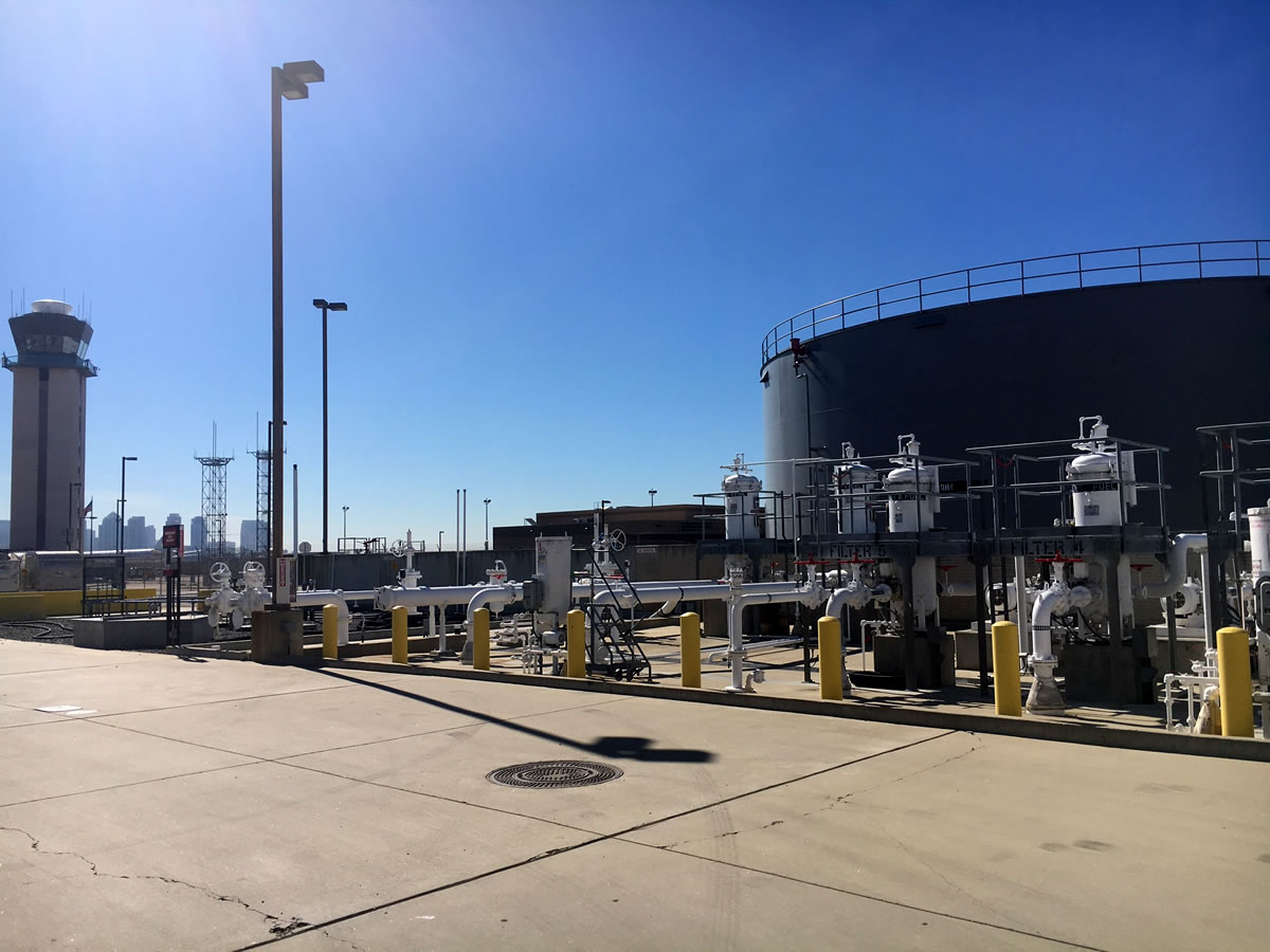 Industrial site with pipeline valves and meters on a clear day, featuring a large storage tank and a fuel offload island in the background.