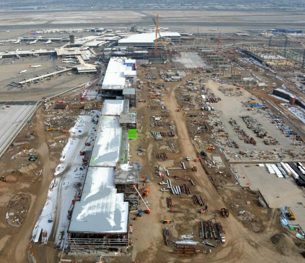 Aerial view of Salt Lake City International Airport expansion site with active construction, featuring equipment and partially constructed buildings, set against a backdrop of snowy mountains.