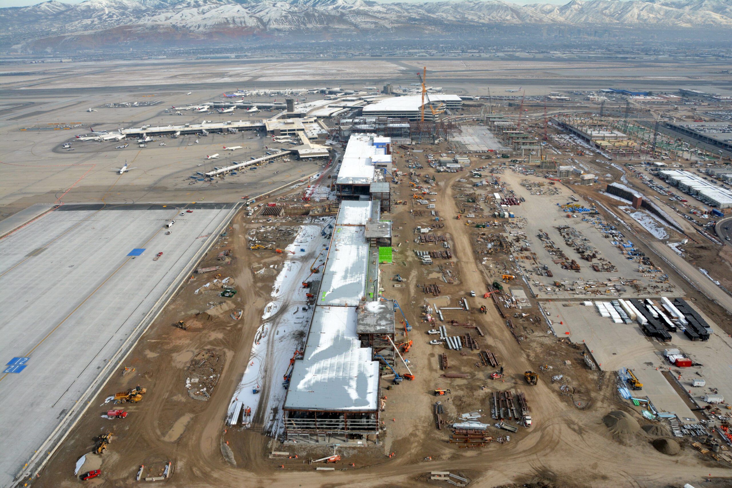 Aerial view of Salt Lake City International Airport expansion site with active construction, featuring equipment and partially constructed buildings, set against a backdrop of snowy mountains.