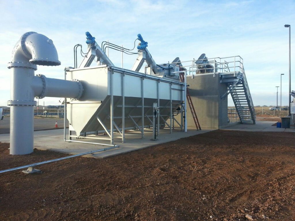 Industrial water treatment facility with large pipes and tanks, equipped with ladders and railing under a clear sky.