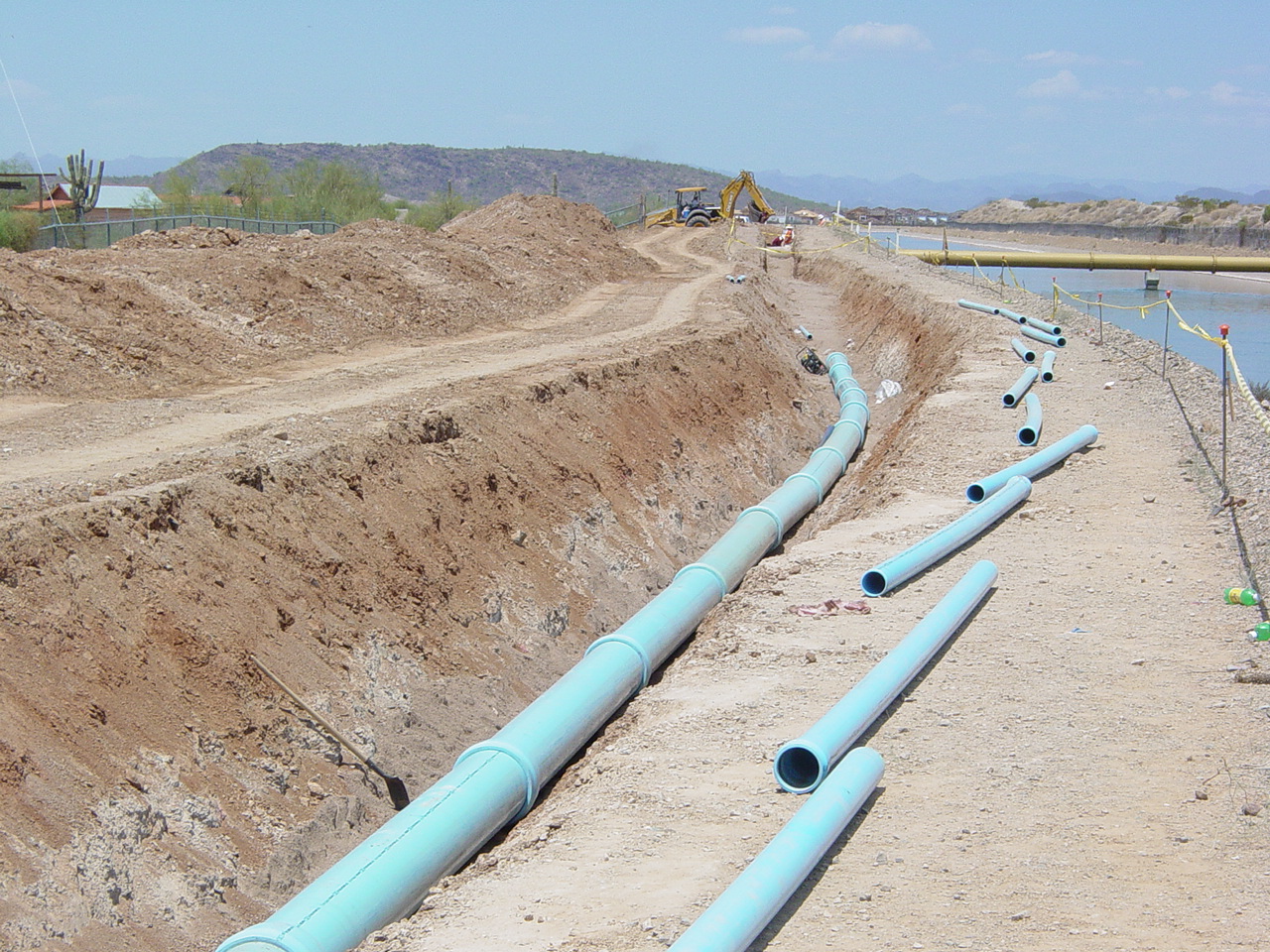A construction site in Phoenix, AZ with large blue pipes laid out along a dug trench for the CAP Dual Waterlines project, featuring excavation machinery and hills in the background.