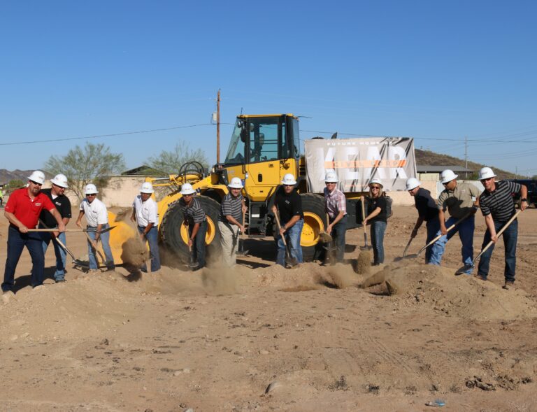 Group of construction workers with shovels breaking ground at a construction site, with a yellow bulldozer in the background about to start work.